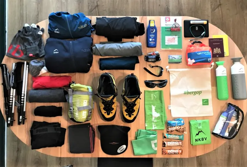 Wandeluitrusting uitgestald op een tafel, waaronder kleding, wandelschoenen, handschoenen, wandelstokken, een zonnebril, toiletartikelen, snacks, waterflessen, kaarten, een paspoort, een EHBO-doos en diverse outdooraccessoires.